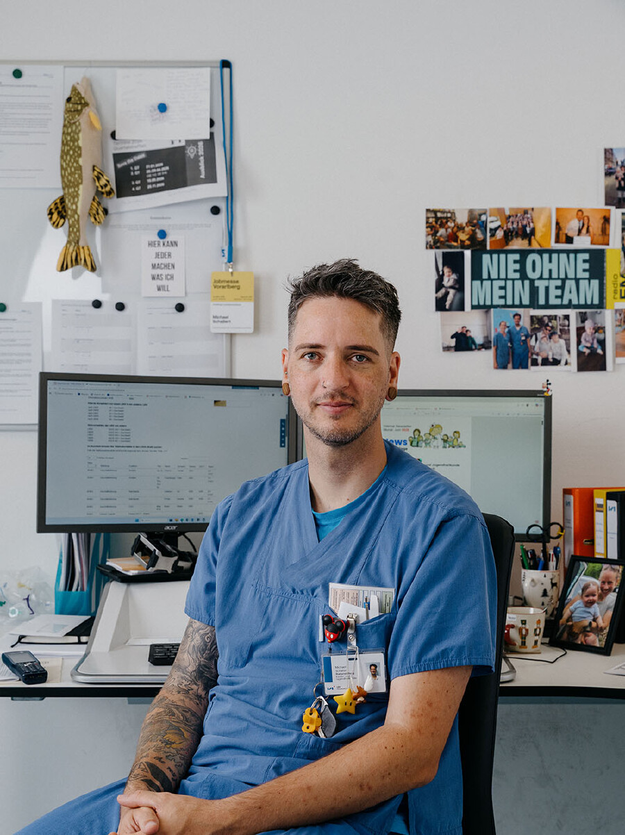 Ein Mann mit kurzen, dunkelbraunen, an den Seiten rasierten Haaren und einem leichten Drei-Tage-Bart sitzt an einem Schreibtisch im B&uuml;ro einer Kinderstation und blickt ruhig in die Kamera. Er tr&auml;gt ein hellblaues medizinisches Kasack-Oberteil, an dessen Brusttasche Stifte, ein Namensschild sowie verspielte Anh&auml;nger (darunter eine Mickey-Mouse-Figur und ein gelber Nintendo-Stern) befestigt sind. Sein rechter Unterarm ist gro&szlig;fl&auml;chig t&auml;towiert. Im Hintergrund stehen zwei Computermonitore mit ge&ouml;ffneten Programmen. Die wei&szlig;e Wand dahinter ist mit einer Magnettafel voller Notizen, einem Stofffisch, privaten Fotos und einer Postkarte mit der Aufschrift &bdquo;NIE OHNE MEIN TEAM&ldquo; dekoriert.