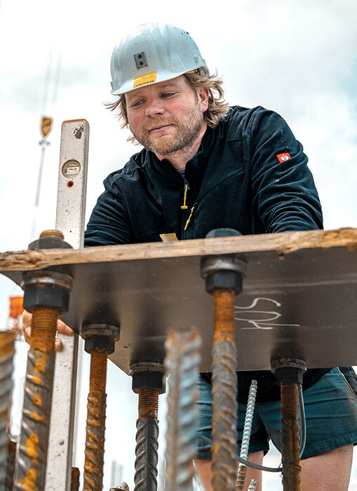 Ein Mann mit hellen Haaren und einem Bart, der als Hochbauer arbeitet, ist von Nahem aus einer tiefen Perspektive zu sehen. Er blickt konzentriert nach unten auf eine Konstruktion aus dicken Gewindestangen und einer Metallplatte, an der er arbeitet. In seiner rechten Hand h&auml;lt er eine silberne Wasserwaage vertikal an die Konstruktion. Er tr&auml;gt eine schwarze Fleecejacke, kurze Arbeitshosen und einen wei&szlig;en Schutzhelm mit einem gelben Logo. Im unscharfen Vordergrund ragen Bewehrungsst&auml;hle auf. Im hellen, bew&ouml;lkten Hintergrund ragt auf der linken Seite ein gro&szlig;er, gelber Turmdrehkran in die H&ouml;he.