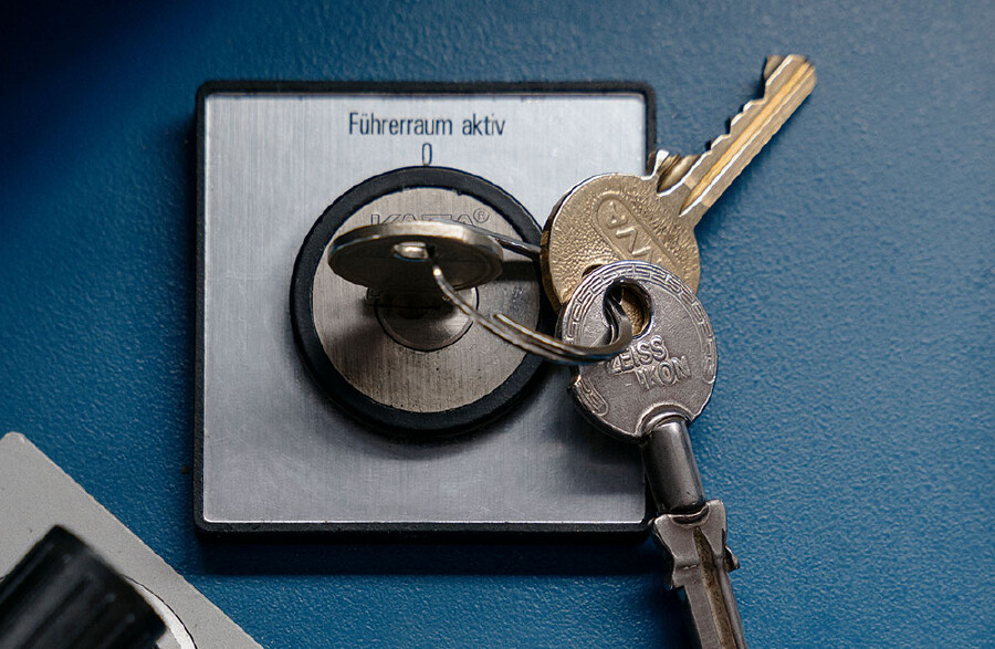 Nahaufnahme eines Schl&uuml;sselbundes, der in einem Schloss auf einer blauen Armaturentafel steckt. &Uuml;ber dem Schloss steht der Text &bdquo;F&uuml;hrerraum aktiv&ldquo;. Ein silberner Schl&uuml;ssel steckt direkt im Zylinder, w&auml;hrend zwei weitere Schl&uuml;ssel (einer goldfarben, einer silberfarben mit der Pr&auml;gung &bdquo;Zeiss Ikon&ldquo;) an einem Ring herabh&auml;ngen.
