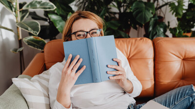 Eine Frau mit Brille sitzt auf einem braunen Ledersofa und h&auml;lt sich schmunzelnd ein ge&ouml;ffnetes, blaues Buch vor die untere Gesichtsh&auml;lfte. Im Hintergrund stehen mehrere gr&uuml;ne Zimmerpflanzen.