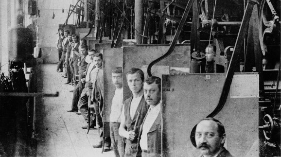 Eine historische Schwarz-Wei&szlig;-Aufnahme aus dem Inneren einer Fabrik. Eine lange Reihe von Arbeitern steht hintereinander an ihren jeweiligen Arbeitsstationen, die durch trennende Platten voneinander abgeteilt sind. Die M&auml;nner blicken alle in Richtung der Kamera. Im Vordergrund ist das Gesicht eines Mannes mit Schnurrbart angeschnitten zu sehen. Entlang der Decke und W&auml;nde verlaufen komplexe mechanische Vorrichtungen, Riemen und Maschinenteile. Der Boden besteht aus Holzdielen.