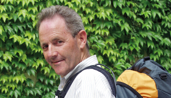 Alt-AK-Pr&auml;sident Josef Fink dreht sich l&auml;chelnd zur Kamera um, einen Rucksack auf dem R&uuml;cken. Der Rucksack steht symbolisch f&uuml;r das von ihm mitinitiierte Abfertigungsmodell &sbquo;Abfertigung neu', das Arbeitnehmerinnen und Arbeitnehmer wie einen mitwachsenden Rucksack durchs Berufsleben begleitet. Im Hintergrund eine gr&uuml;ne Blattwand.