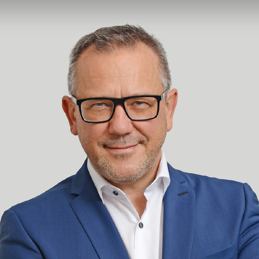 Ein Portr&auml;tfoto eines l&auml;chelnden, mittelalten Mannes mit grauem Haar, Bartstoppeln und einer Brille mit schwarzem Rahmen. Er tr&auml;gt einen blauen Anzug und ein wei&szlig;es Hemd, seine Arme sind verschr&auml;nkt und er tr&auml;gt eine Armbanduhr am linken Handgelenk.
