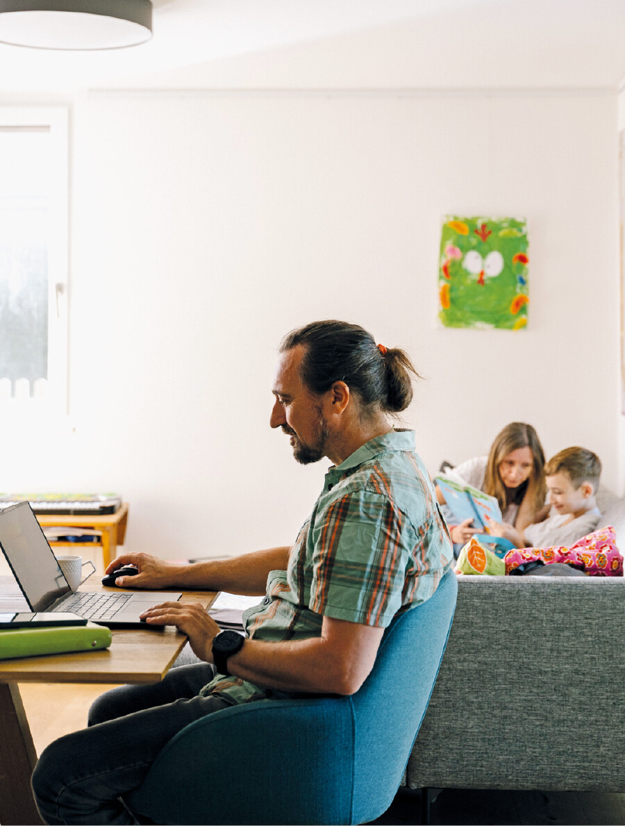 Ein Mann mit Pferdeschwanz und kariertem Kurzarmhemd sitzt im Profil an einem Holztisch und arbeitet an einem Laptop. Im Hintergrund liest eine Frau mit einem Kind auf dem Sofa in einem Buch. An der hellen Wand h&auml;ngt ein buntes Kinderbild.