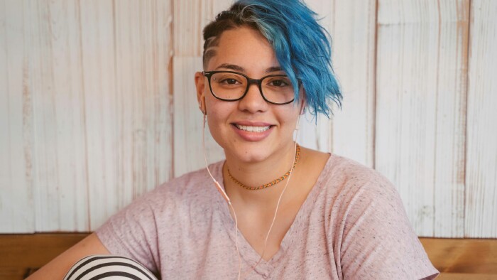 Eine Frau mit blauen Haaren und einer Brille sitzt auf einer Bank und blickt in die Kamera.