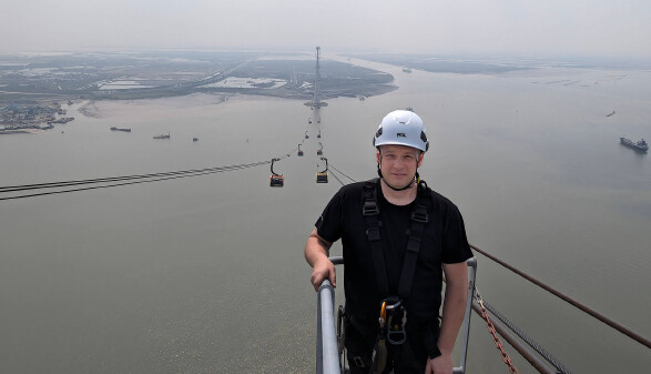 Seilbahntechniker steht auf der Plattform an der Spitze einer Seilbahnstütze, unter ihm sieht man das Meer und im Hintergrund die Seilbahn, die bis zum Festland am Horizont reicht.