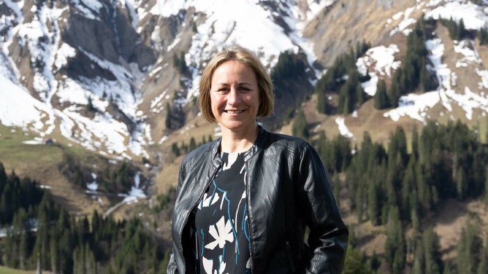 Eine Frau mit halblangen, blonden Haaren, Angela Schwarzmann, lächelt in die Kamera. Im Hintergrund sieht man ein beeindruckendes Bergpanorama, auf dem teils noch Schnee liegt.