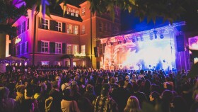 Ein gro&szlig;e Menschenmenge steht vor einer beleuchteten B&uuml;hne, auf der eine Band gerade ein Konzert gibt.