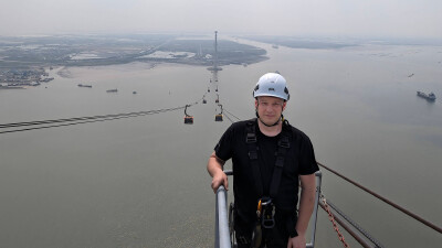 Seilbahntechniker steht auf der Plattform an der Spitze einer Seilbahnstütze, unter ihm sieht man das Meer und im Hintergrund die Seilbahn, die bis zum Festland am Horizont reicht.