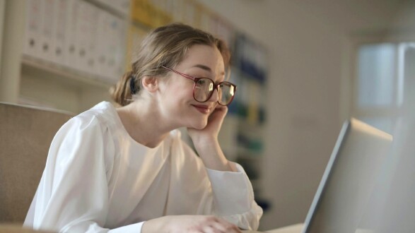 Eien Frau mit Brille, die zufrieden am Schreibtisch sitzt und in einen Bildschirm schaut