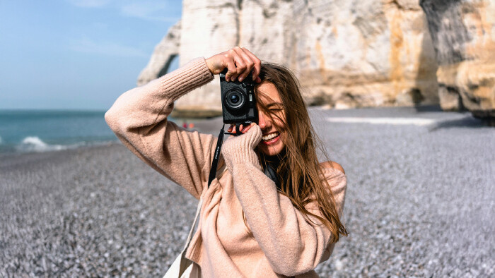 Eine Frau steht mit einer Fotokamera am Strand und lacht.