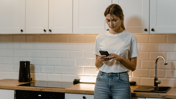 Eine junge Frau in wei&szlig;em T-Shirt und Jeans steht in einer hellen K&uuml;che. Sie h&auml;lt ihr Smartphone mit beiden H&auml;nden und schaut mit ernstem, skeptischem Blick auf den Bildschirm.