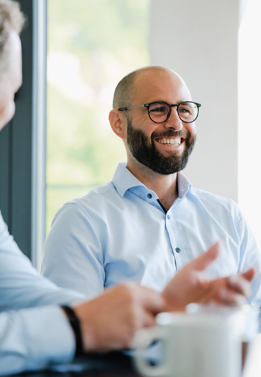 Portr&auml;t eines l&auml;chelnden Manns mit Glatze, dunklem Vollbart, schwarzer Brille und hellblauem Hemd. Er sitzt an einem Tisch und blickt aufmerksam zu seinem Gespr&auml;chspartner, dessen H&auml;nde im unscharfen Vordergrund zu sehen sind. Im Hintergrund ist ein helles Fenster mit Blick ins Gruene.