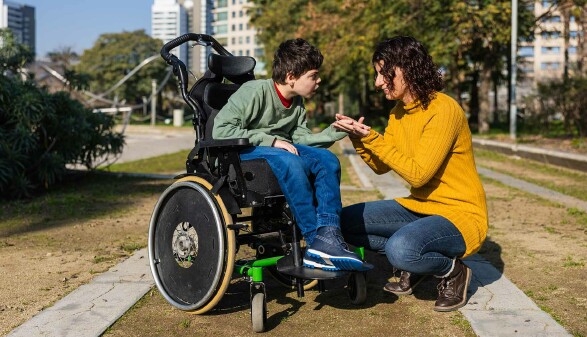 Eine Frau kniet vor einem Jungen im Rollstuhl und hält seine Hand. Im Hintergrund ist ein Park zu sehen, die Sonne scheint.