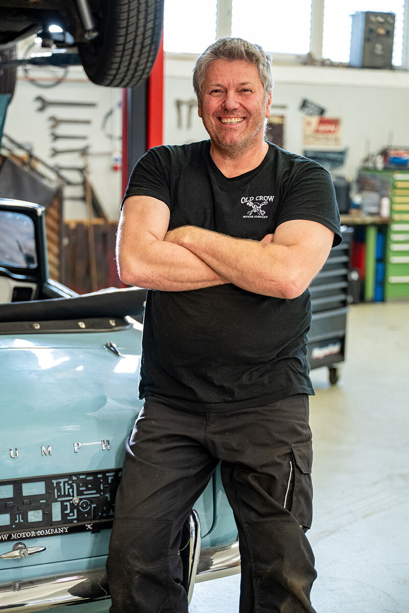 Werner Metzler steht l&auml;chelnd mit verschr&auml;nkten Armen vor einem hellblauen Triumph-Cabriolet in seiner Werkstatt. Er tr&auml;gt ein schwarzes T-Shirt mit dem Logo seiner Oldtimer-Werkstatt Old Crow Motor Company.