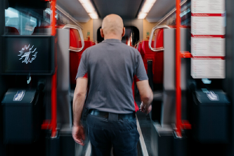 R&uuml;ckansicht von Holger Hansen, einem glatzk&ouml;pfigen Mann in Dienstkleidung (graues Poloshirt mit roten Akzenten und dunkle Hose), der durch den Gang eines modern gestalteten Zuges geht. Die Umgebung mit roten Sitzen und Haltestangen wirkt durch eine Bewegungsunsch&auml;rfe dynamisch, w&auml;hrend die Person im Zentrum scharf fokussiert ist.