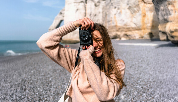 Eine Frau steht mit einer Fotokamera am Strand und lacht.