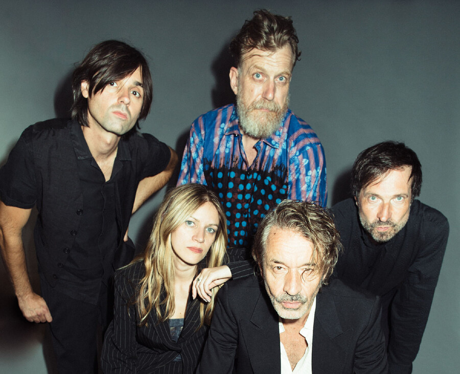 Ein Gruppenportr&auml;t der f&uuml;nfk&ouml;pfigen Band Naked Lunch vor einem schlichten grauen Hintergrund. F&uuml;nf Bandmitglieder schauen in die Kamera. In der Mitte befindet sich eine Frau mit langen blonden Haaren, umgeben von vier M&auml;nnern. Einer der M&auml;nner hat einen markanten Bart und tr&auml;gt ein auff&auml;lliges blaues Musterhemd, w&auml;hrend die anderen M&auml;nner &uuml;berwiegend schwarze Kleidung tragen. Die Bandmitglieder schauen mit ernsten Ausdr&uuml;cken in die Kamera.