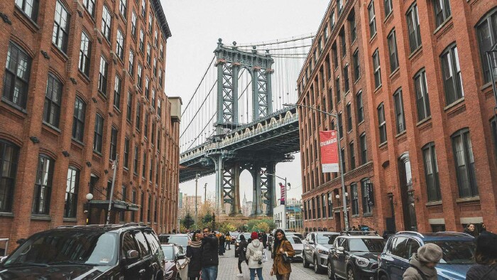 Mehrere Menschen stehen auf einer Straße in New York, im Hintergrund sieht man die berühmte Brooklyn Bridge.