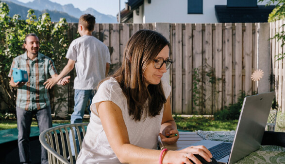 Adrijana K&uuml;nzle sitzt an einem Tisch im Garten und arbeitet an einem Laptop. Im Hintergrund stehen Andreas und David K&uuml;nzle auf dem Rasen vor einem Holzzaun und unterhalten sich. Im Hintergrund erstreckt sich eine Bergkulisse.