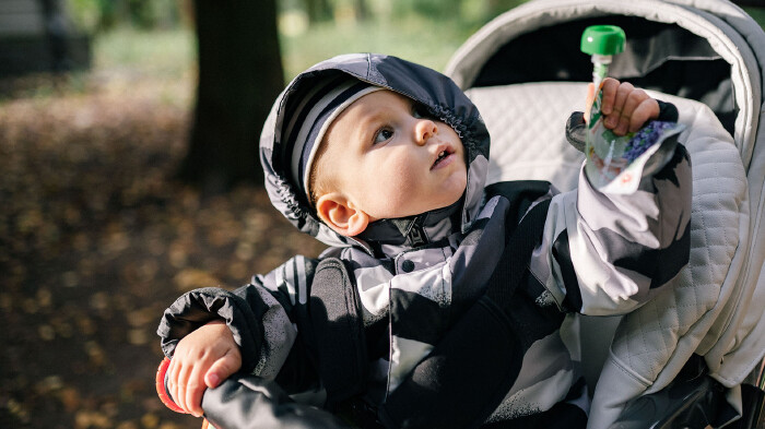Ein Baby sitzt angeschnallt in einem Kinderwagen und h&auml;lt eine Trinkflasche in der Hand. Der Kinderwagen steht drau&szlig;en in einer gr&uuml;nen Umgebung.