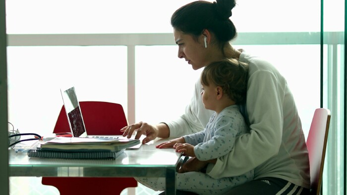 Eine Frau, gekleidet in ein wei&szlig;es Sweatshirt und eine graue Jogginghose, sitzt an einem wei&szlig;en Schreibtisch und schaut konzentriert auf einen Laptop. Ihre Haare sind zu einem Knoten hochgebunden und sie tr&auml;gt wei&szlig;e Ohrh&ouml;rer. Auf ihrem Scho&szlig; sitzt ein kleines Kind in einem hellblauen Strampler. Das Kind blickt ebenfalls auf den Laptop. Auf dem Tisch liegen ein Notebook und Stifte. Der Hintergrund ist hell, mit einem gro&szlig;en Fenster und wei&szlig;en Fensterrahmen.