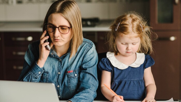 Eine Mutter sitzt am Tisch, telefoniert und blickt auf einen Laptop, während ihre Tochter daneben zeichnet. Die Szene spielt in einer Küche und zeigt eine Alltagssituation zwischen Familienleben und beruflicher Verantwortung.