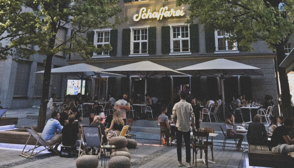 Abendliche Au&szlig;enansicht des Geb&auml;udes der Schaffarei im AK Quartier. Im geschm&uuml;ckten Au&szlig;enbereich sitzen zahlreiche Personen an Tischen unter gro&szlig;en wei&szlig;en Schirmen sowie auf Liegest&uuml;hlen. &Uuml;ber der Fassade leuchtet der Schriftzug &bdquo;Schaffarei&ldquo; in gelber Neonschrift.