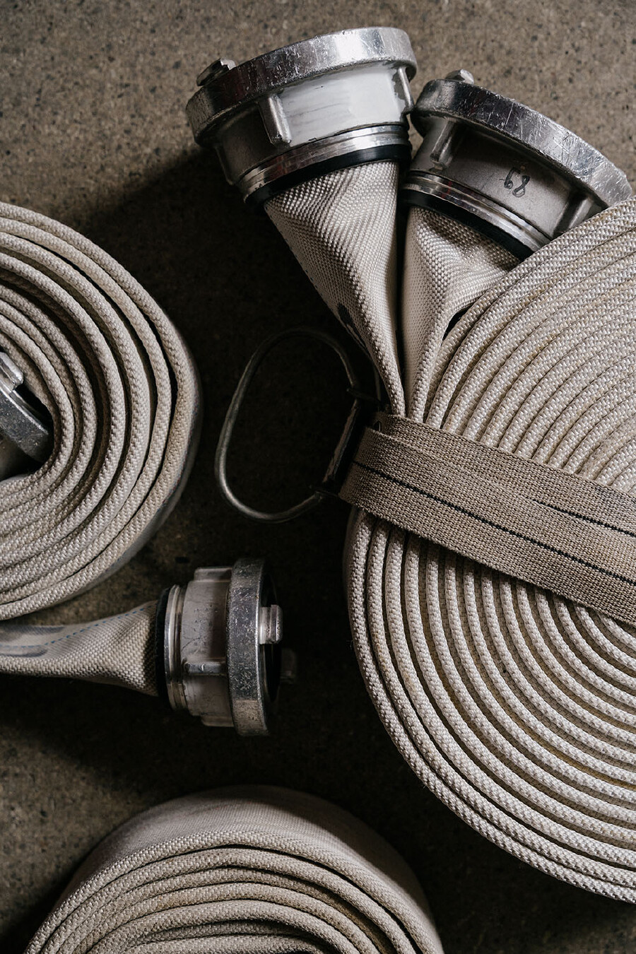 Eine Draufsicht auf mehrere beige Gewebe-Feuerwehrschl&auml;uche, die auf einem Betonboden liegen. Auf der rechten Seite sind zwei Schl&auml;uche mit gro&szlig;en, metallischen Storz-Kupplungen parallel aufgerollt und werden durch einen dicken, braunen Textilgurt mit einem Metall-D-Ring zusammengehalten. Links daneben und darunter sind ein weiterer sauber aufgerollter Schlauch sowie das Ende eines flach liegenden Schlauchs mit einer einzelnen Metallkupplung zu sehen. Am unteren Bildrand ist ein weiterer Schlauchstapel im Anschnitt erkennbar. Die Schl&auml;uche weisen leichte Gebrauchsspuren auf.