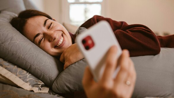 Eine Frau liegt lächelnd im Bett und hält ein Handy in der Hand. 