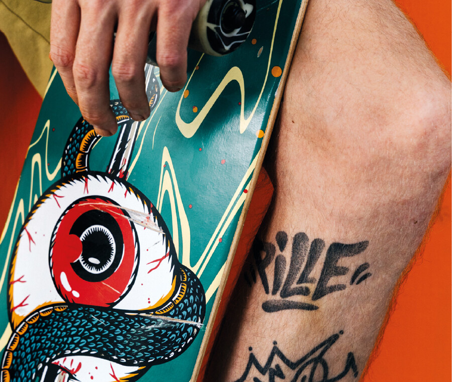 Nahaufnahme einer Hand, die ein gr&uuml;nes Skateboard mit einer Augen-Grafik h&auml;lt. Daneben ist das Bein einer Person mit Tattoos zu sehen, unter anderem mit dem Schriftzug &bdquo;PILLE&ldquo;. Der Hintergrund ist orange.