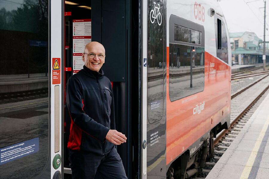 Holger Hansen steht l&auml;chelnd in der offenen T&uuml;r eines &Ouml;BB Cityjet-Zuges an einem Bahnsteig. Er tr&auml;gt eine dunkelblaue Fleecejacke mit roten Eins&auml;tzen und eine Brille. Der rot-wei&szlig;-graue Zug ist im Profil zu sehen, im Hintergrund erkennt man die Gleise und das Bahnhofsgel&auml;nde.